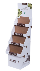 A white cardboard display stand with four shelves, each labeled "Aloha." The top section reads, "Plant-based goodness is our specialty" with sharp green leaf illustrations. The base features a brown graphic of palm trees and the brand name "Aloha." The shelves are empty, awaiting your products' packaging.