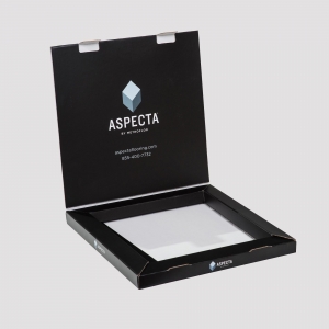 An open black cardboard box with a lid displaying the text "ASPECTA BY METROFLOR" along with a logo. Inside, there is a transparent window showcasing the content of the packaging beneath. Contact information is printed below the logo. The background is plain light gray.