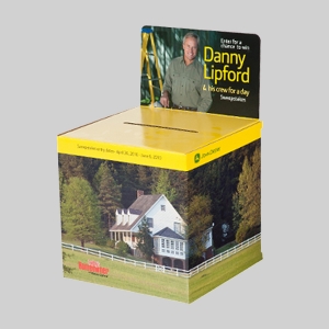 A display box with a photo of a white house surrounded by trees on the front. The top has a slot for entries and a sign featuring graphics of a man in work attire. Text on the sign reads: "Enter for a chance to win Danny Lipford & his crew for a day Sweepstakes.