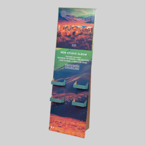 A tall, narrow standee display features album promotion graphics. The packaging highlights a new studio album with text promoting songs like "Scandal, Be Care, and Relentless," and mentions a "Deluxe Edition." The background shows a colorful desert landscape.