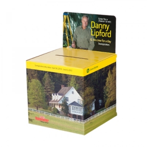 A cardboard contest entry box, adorned with sharp graphics, features an image of a man standing on a ladder and text offering a chance to win a day with "Danny Lipford & his crew." The box is decorated with an image of a white house with a porch, surrounded by greenery and trees.