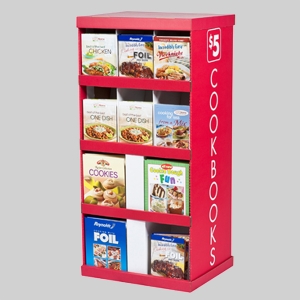 A red display shelf labeled "Cookbooks $5" features various cookbooks and cooking-related items on six shelves. The items, adorned with sharp graphics, include books on chicken recipes, one-dish meals, cookies, and cooking for kids, along with packages of Reynolds Wrap aluminum foil.