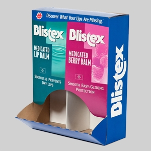 A blue cardboard display box with sharp graphics holds two types of Blistex lip balm. The green packaging on the left is for Medicated Lip Balm, soothing and preventing dry lips. The pink packaging on the right is for Medicated Berry Balm, providing smooth, easy-gliding protection.