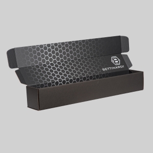 A black cardboard box, resembling a sleek shipping container, is open to reveal a striking hexagonal pattern on its inner surface. The right side of the inner lid features sharp graphics of the "Bettinardi" logo and name. The package is set against a plain, light gray background.