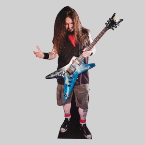 A person with long hair and a beard, wearing a black shirt, cargo shorts, and sneakers, stands holding a blue electric guitar. The person has tattooed legs and wears red socks. They lean casually against a shipping container in the plain gray background.