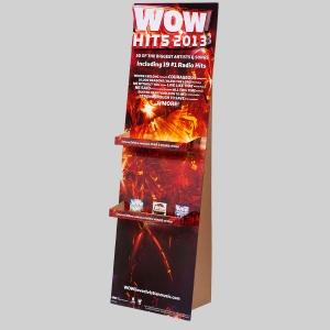 A cardboard display stand for WOW Hits 2013 is shown. It has a fiery red and yellow background with text promoting the music compilation, mentioning 30 of the biggest artists and songs, including 19 #1 Radio Hits. The attractively packaged bottom shelf holds various CD cases.