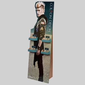 A promotional cardboard display stand featuring a person with blond hair in a jacket adorned with patches. The stand, labeled "Colton Dixon," has four shelves holding small packages and is set against a blurred background with a dry, cracked earth floor.