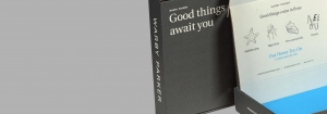 A close-up of an open Warby Parker subscription box is shown. The left side of the package displays the text "Good things await you." Inside the lid are sharp graphics and text, including "Good things come in fives" with icons representing sunglasses, eyeglasses, and a home try-on kit.