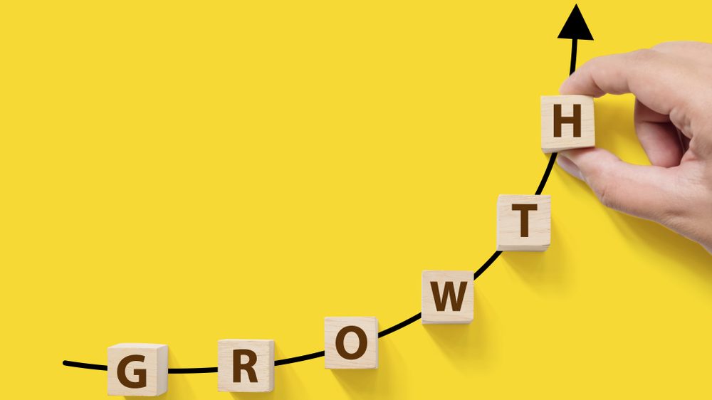 A hand places a wooden block with the letter "H" at the end of a curved, upward-pointing black arrow. Sharp graphics adorn other blocks that spell "GROWTH" on a bright yellow background, symbolizing the concept of growth and upward progress.