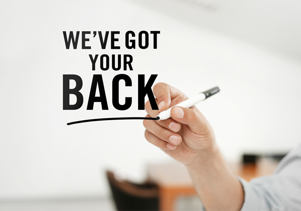 A close-up of a hand holding a black marker pen, drawing a horizontal line underneath the text "WE'VE GOT YOUR BACK." The background is blurred, with indistinct furniture and sharp graphics suggesting an office environment.