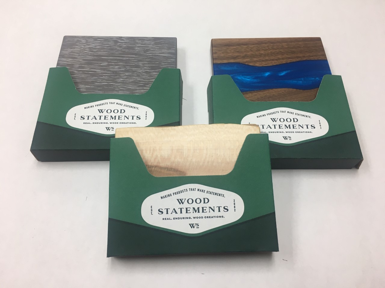 Three green packages labeled "Wood Statements" are displayed. Each package contains a different type of wood sample: one greyish wood, one with a blue epoxy resin design, and one light-colored wood with a subtle pattern. The packaging exudes elegance, reinforcing the tagline "real designers, wood creations.