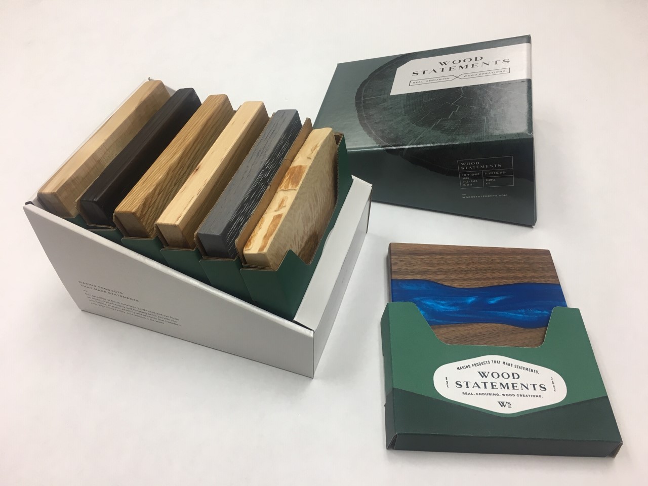 A display of "Wood Statements" samples in a cardboard holder. Various wood samples with different finishes and colors are visible. One green and white subscription box shows a sample with a striking blue resin inlay. Another box is partially open, revealing more wood samples with sharp graphics.