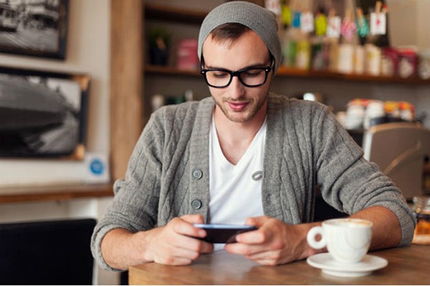 A man with glasses and a gray beanie sits at a wooden table in a cozy cafe, looking at his smartphone. He is wearing a gray cardigan over a white t-shirt. A cup of coffee sits on the table in front of him, next to a small cardboard box, and the cafe background is slightly blurred.