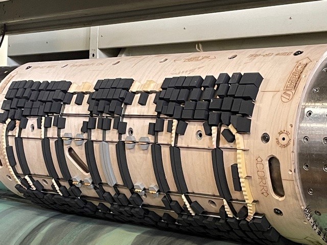 Close-up view of an industrial machine part with a cylindrical drum featuring various black and beige rubber elements, likely for printing or stamping purposes. The surface includes detailed engravings and markings with symbols and text, ideal for creating graphics on packaging materials.