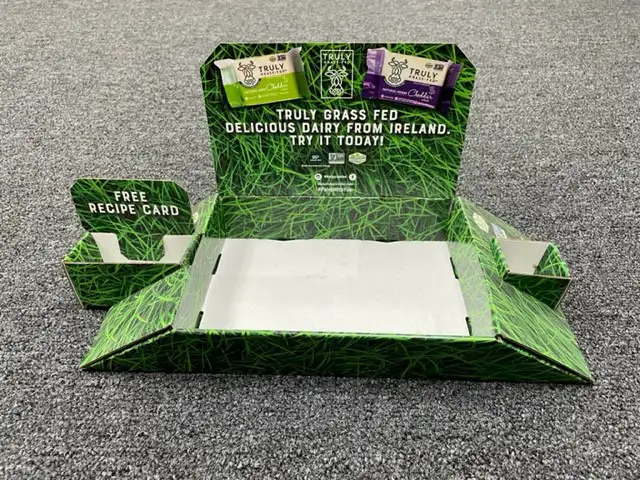 A green, grass-themed cardboard packaging on a gray carpet showcases Truly Grass Fed dairy products from Ireland. The package includes labels of butter variations at the top and a section for free recipe cards on the left. Text reads, "DELICIOUS DAIRY FROM IRELAND, TRY IT TODAY!