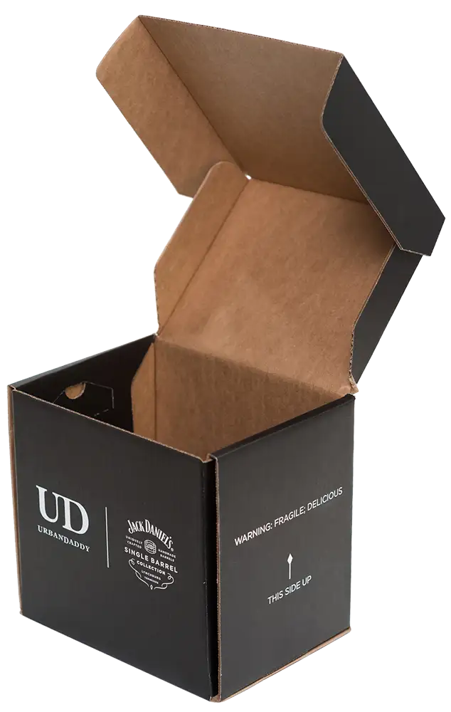 A black and brown cardboard box is open, displaying its interior. The package features the UrbanDaddy logo, a Jack Daniel's Single Barrel Select label, and the warning "FRAGILE, DELICIOUS" with an arrow indicating "THIS SIDE UP" along with sharp graphics that emphasize its premium content.