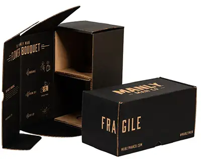 Two black cardboard boxes with the text "MANLY MAN" and "FRAGILE" on the front. One box is open, displaying inner compartments perfect for organized packaging. The other shipping container is closed, positioned horizontally next to the open box.