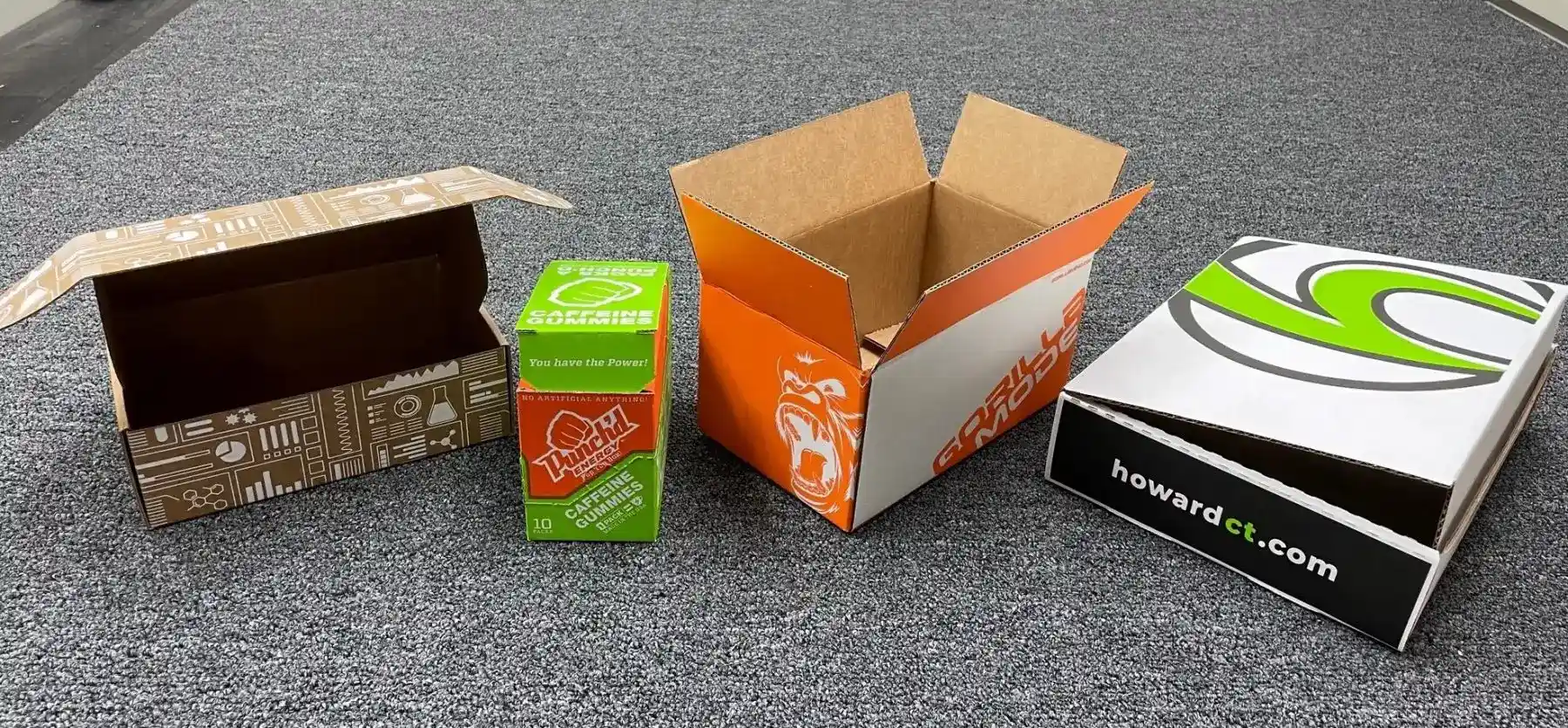 Four different open cardboard boxes are displayed on a gray carpet. From left to right: a brown box with graphics and symbols, a small green package labeled "Ticasi," an orange and white box with the text "Golf," and a white and black branded box with the website "howardt.com.